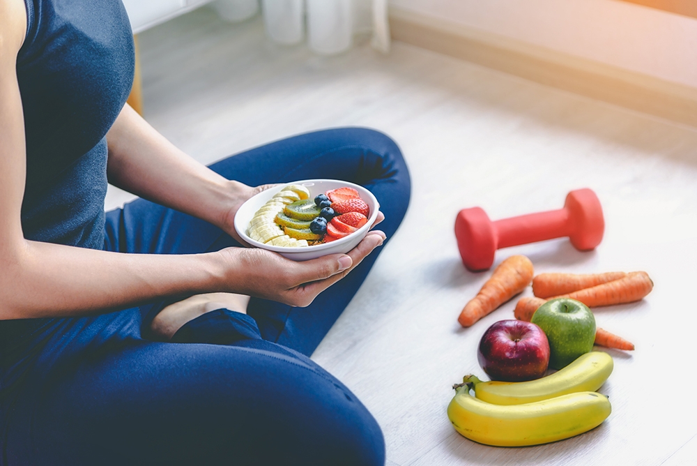 Woman holding fruit salad, next to weights.