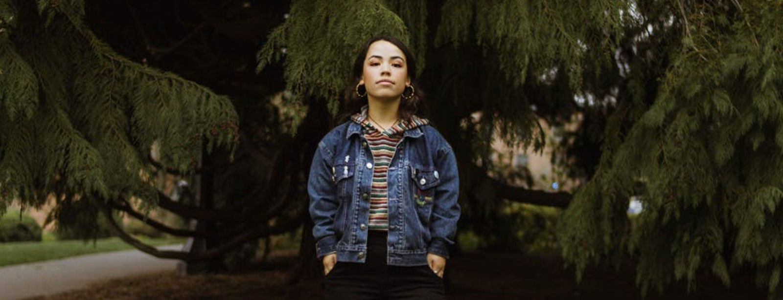 Person in denim jacket standing under trees.