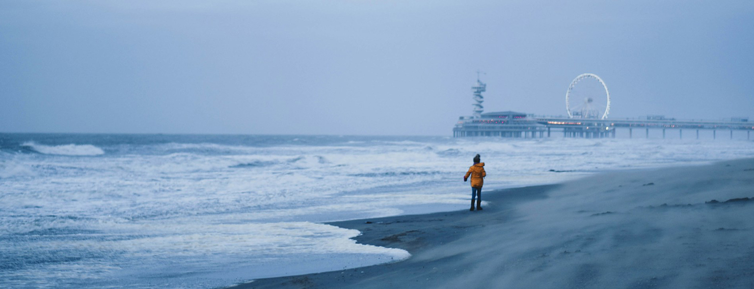 Child walks on a windy beach near a pier.