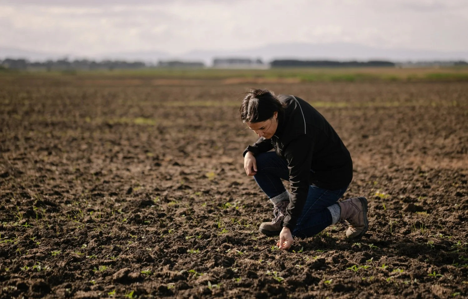 Woman kneels examining soil in a large field.